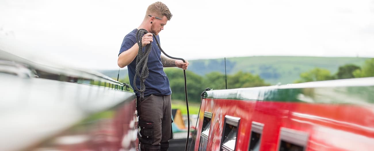 A Snaygill boatyard hand coils a mooring line between two narrowboats at Bradley, with Dales hills beyond